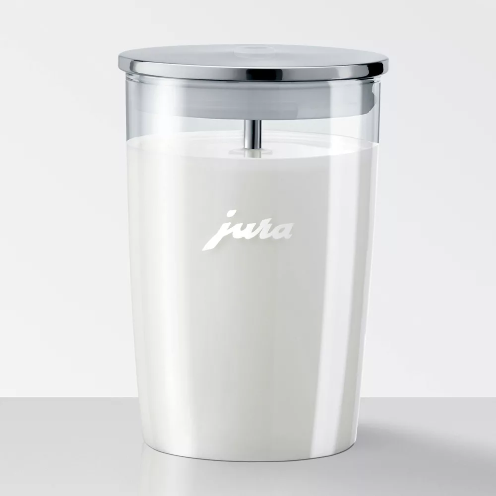 A Jura Glass Milk Container with Lid - 16.9 oz, filled with milk, features a stainless steel lid, internal metal rod, and white jura branding on the front. The container rests on a light surface against a white background. © 2025 1st-line.com