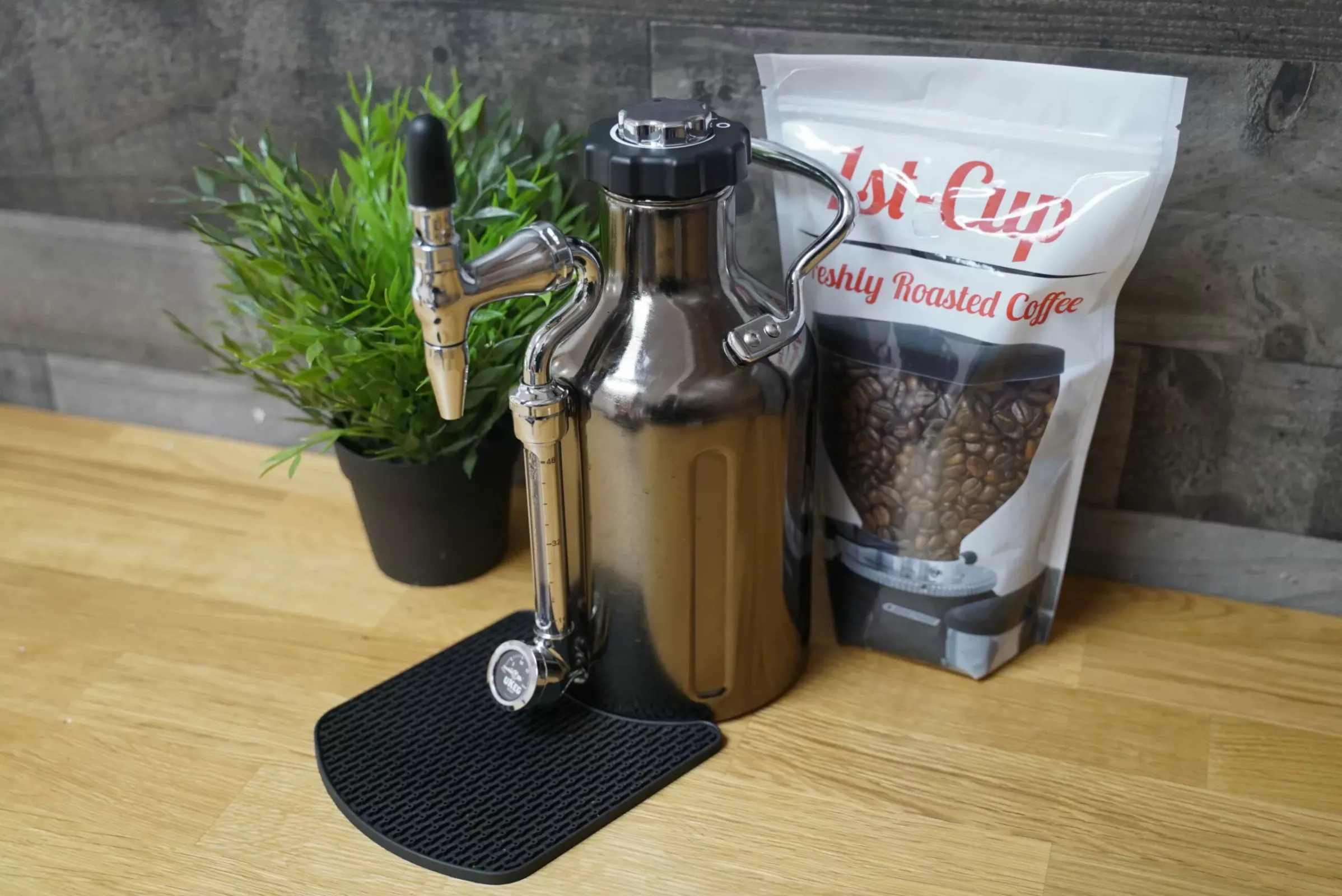 A DISCONTINUED GrowlerWerks uKeg Nitro Cold Brew Coffee Maker with N2O, featuring a tap and pressure gauge, rests on a black mat atop wood near coffee beans and a potted plant, ready for nitro cold brew infusion. © 2025 1st-line.com