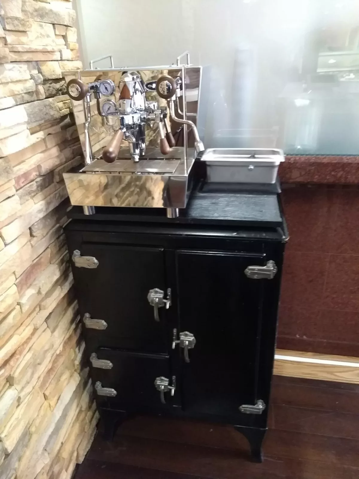 A DISCONTINUED Izzo Valexia HX Espresso Machine sits atop a vintage black icebox with chrome handles, set against a stone wall in a cozy indoor space—a rare find since this model is no longer available. © 2025 1st-line.com