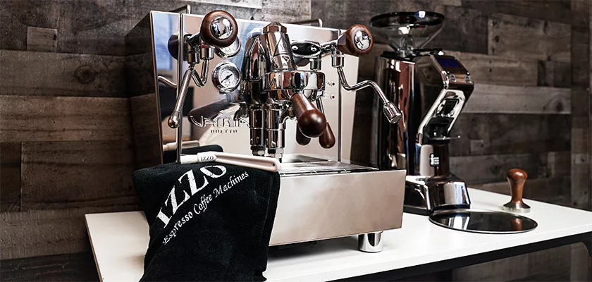 A polished DISCONTINUED Izzo Valexia Duetto II Dual Boiler Espresso Machine with wood accents sits on a white counter beside a grinder, while a black IZZO towel hangs nearby against a backdrop of dark wood paneling. © 2025 1st-line.com