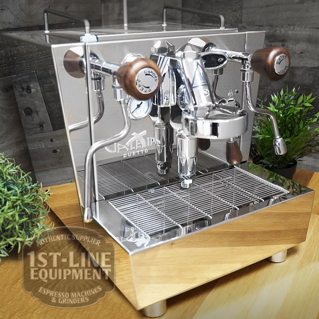 A shiny DISCONTINUED Izzo Valexia Duetto II Dual Boiler Espresso Machine with wooden accents sits on a wood counter between two small green potted plants. “1st-Line Equipment” appears in the lower left corner. © 2025 1st-line.com