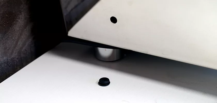 A small black rubber foot rests on a white surface near a modern metallic stand, likely from the DISCONTINUED Izzo Valexia Duetto II Dual Boiler Espresso Machine, beside a slanted white panel with a round hole. © 2025 1st-line.com