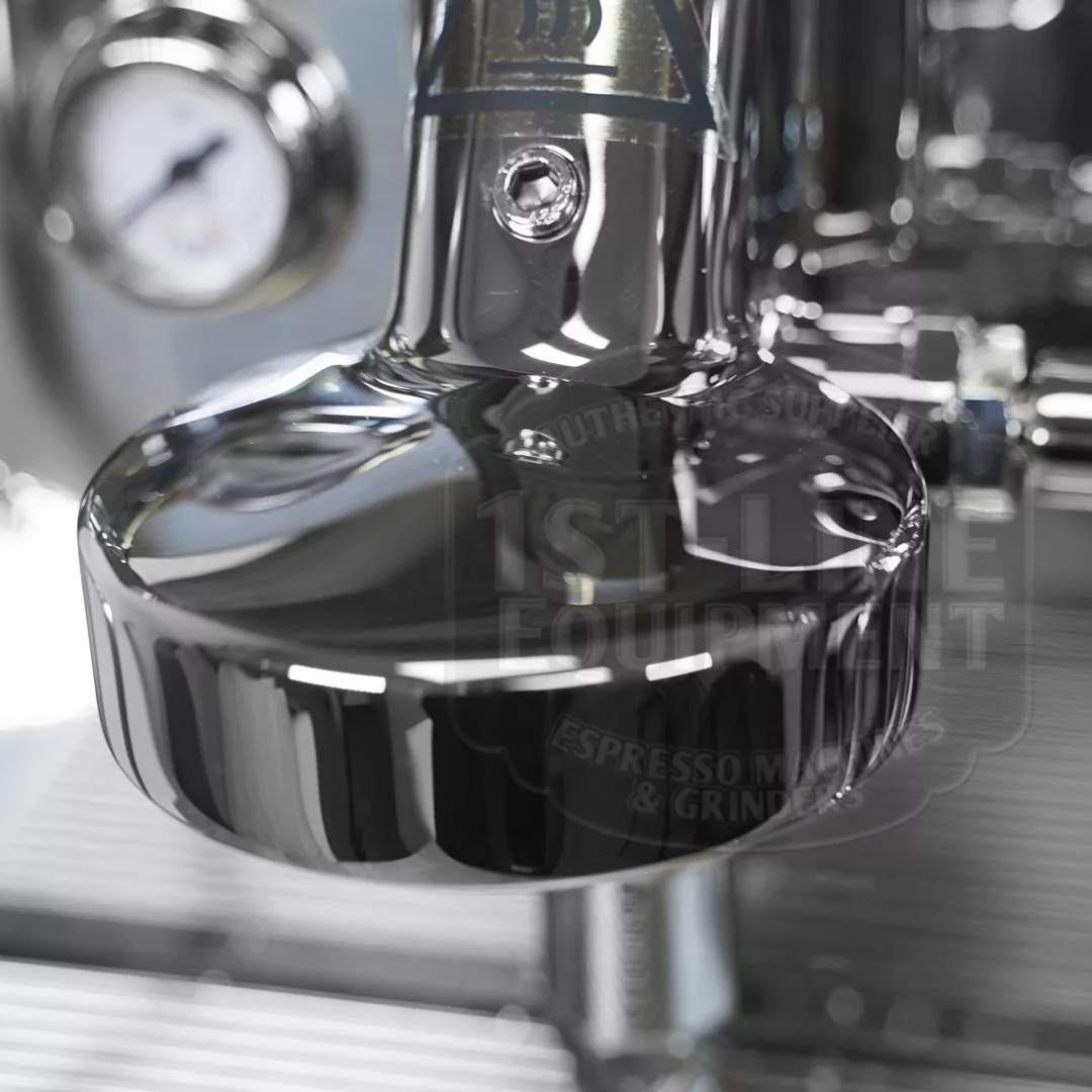 Close-up of a shiny metal espresso machine portafilter with a gauge in the blurred background and reflections visible on the polished surface. © 2025 1st-line.com