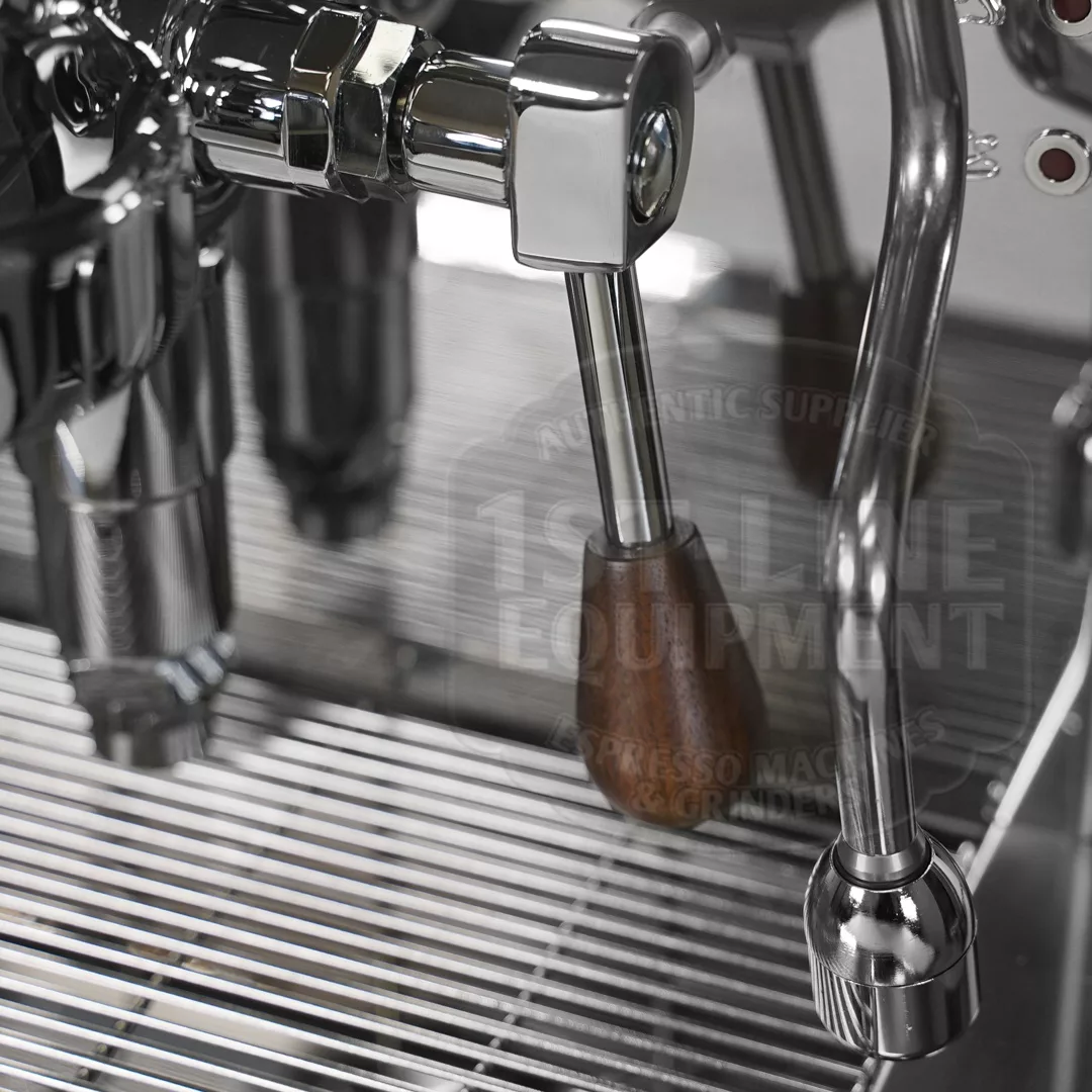 Close-up of a stainless steel espresso machine, focusing on the group head and steam wand with a wooden handle, above a metal drip tray with parallel bars. © 2025 1st-line.com