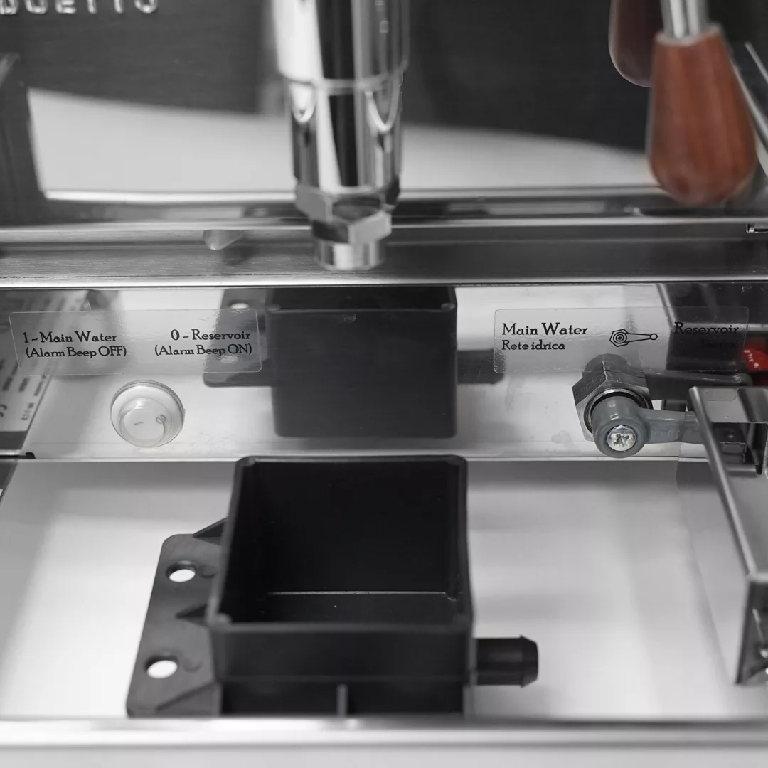 Close-up of internal components on the DISCONTINUED Izzo Valexia Duetto II Dual Boiler Espresso Machine, showing labeled switches for main water or reservoir settings, a black removable tray, and metal and wooden parts. © 2025 1st-line.com