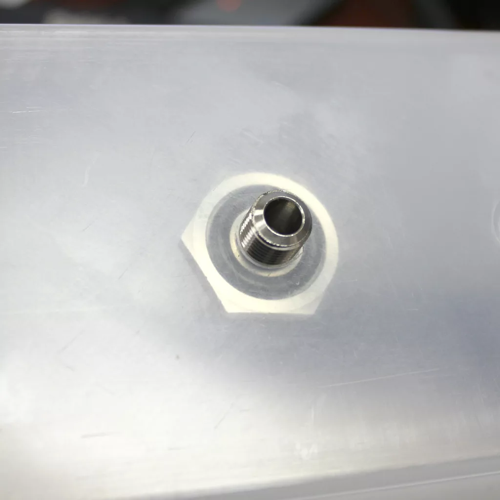 A close-up of a metal threaded water tank adapter installed into a translucent plastic surface, possibly part of a tank or container. © 2025 1st-line.com