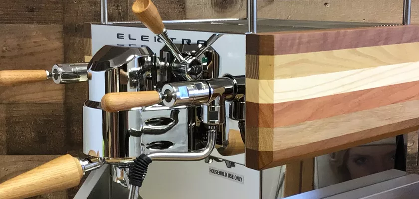 A close-up of the Elektra Verve Dual Boiler Espresso Machine showcases its polished metal finish, wooden handles, and accents, set against a rustic wooden wall for a modern yet warm look. © 2025 1st-line.com