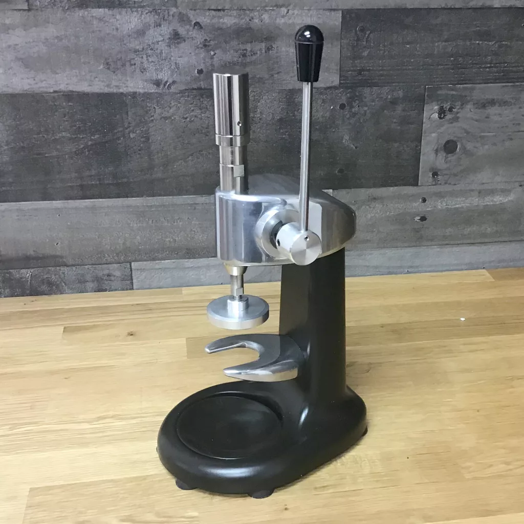 A returned and refurbished Macap CPS Dynamometric Tamper sits on a wooden surface against a wood-paneled wall. © 2025 1st-line.com
