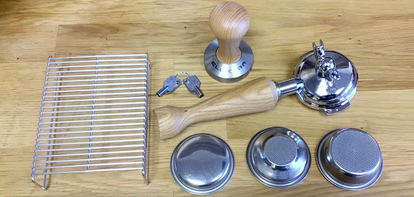 Accessories for the Elektra Verve Dual Boiler Espresso Machine are displayed on a wooden surface, including a metal tamper, two keys, a portafilter with a wooden handle, a metal rack, and three metal filter baskets. © 2025 1st-line.com