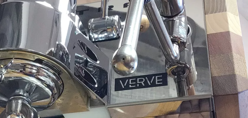 Close-up of the Elektra Verve Dual Boiler Espresso Machine, featuring the VERVE nameplate on its metal body, highlighted by reflective surfaces and visible machine components. © 2025 1st-line.com