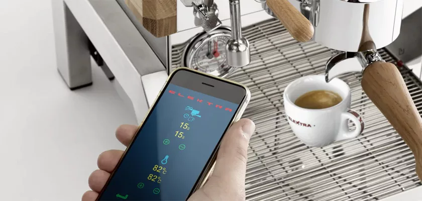Someone holds a smartphone showing Elektra Verve Dual Boiler Espresso Machine settings in front of the machine while it brews espresso into a white cup. © 2025 1st-line.com