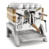 The Elektra Verve Dual Boiler Espresso Machine features a polished chrome finish, wooden handles and side panels, a top cup rack with white cups, and a front pressure gauge showcasing its dual boiler performance. © 2025 1st-line.com