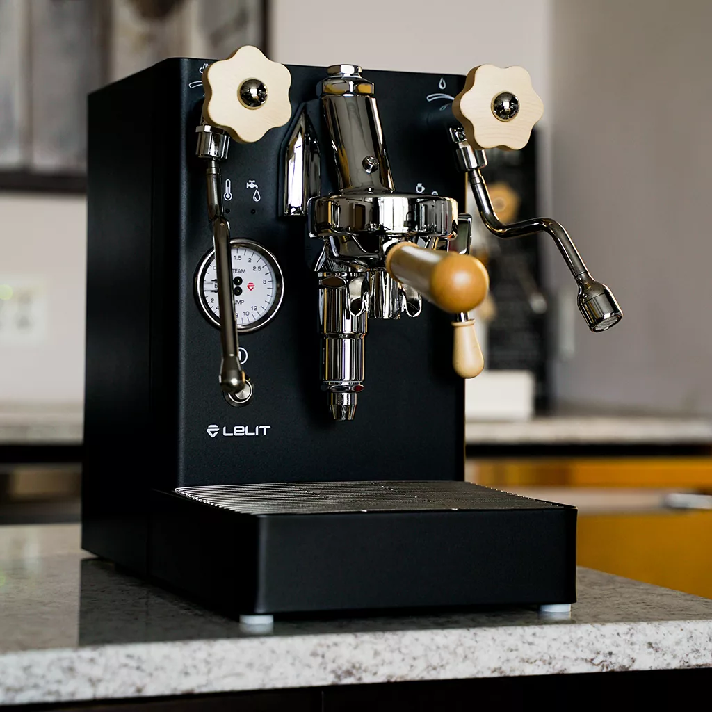 A black Lelit Mara X V2 HX Espresso Machine with chrome and wooden accents sits on a marble counter, showcasing dual handles, a steam wand, and a pressure gauge. © 2025 1st-line.com