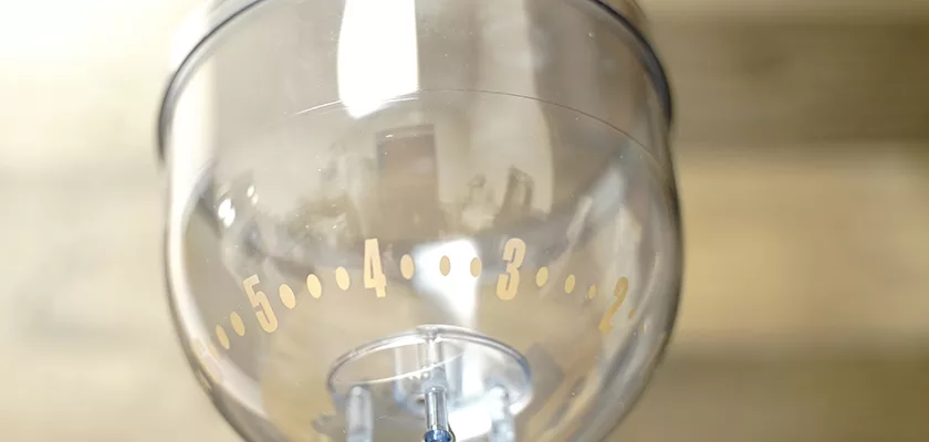 A close-up of a transparent container with markings from 2 to 5, likely part of the discontinued Lelit PL043MMI Fred Espresso Coffee Grinder, set against a blurred background. © 2025 1st-line.com