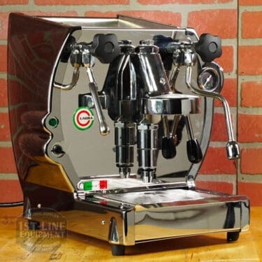 A Like NEW La Nuova Era Cuadra Espresso Machine - V3 Chrome Color (C105) with dual group heads, steam wands, control knobs, and a pressure gauge sits on a counter in front of a red brick wall, displaying the LiaDia brand name. © 2025 1st-line.com