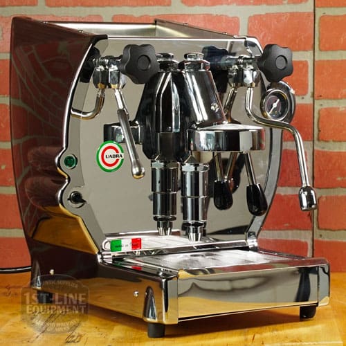 A Like NEW La Nuova Era Cuadra Espresso Machine - V3 Chrome Color (C105) with dual group heads, steam wands, control knobs, and a pressure gauge sits on a counter in front of a red brick wall, displaying the LiaDia brand name. © 2025 1st-line.com