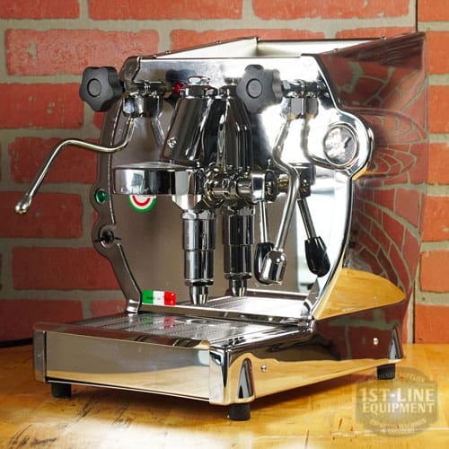 A Like NEW- La Nuova Era Cuadra Espresso Machine - V3 Chrome Color (C105) with dual spouts, steam wand, gauges, and control knobs sits on a wooden counter by a red brick wall, its chrome surface reflecting the surroundings. © 2025 1st-line.com