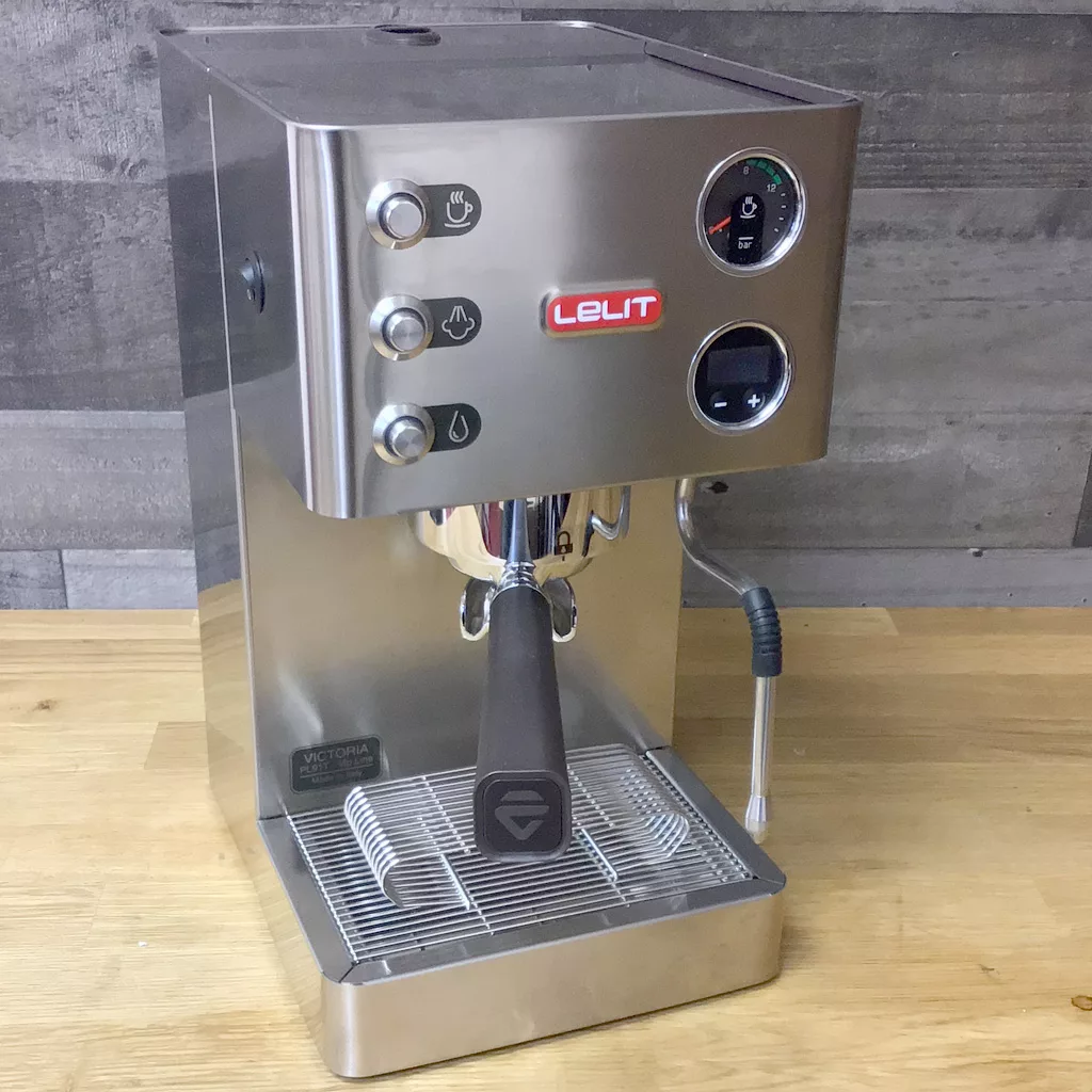 A returned & refurbished Lelit Victoria PL91T PID Single Boiler Espresso Machine with control buttons, pressure gauge, and steam wand sits on a wooden countertop against a gray tile wall. © 2025 1st-line.com