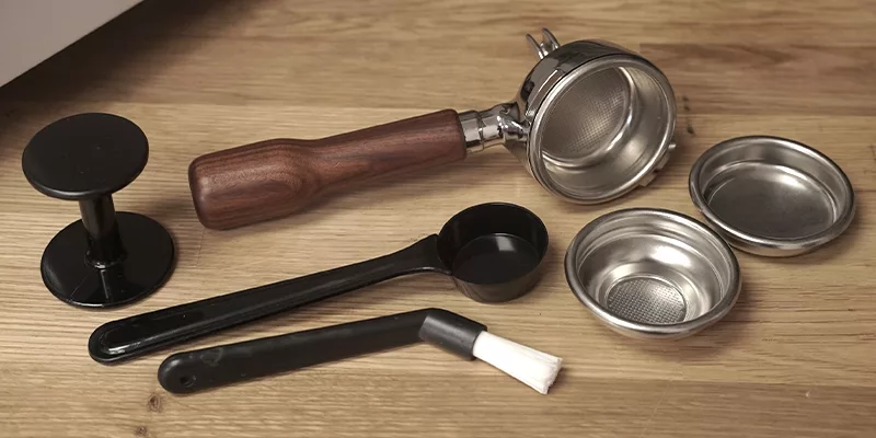 A set of espresso machine accessories on a wooden surface—including a portafilter, filter baskets, tamper, scoop, and cleaning brush—perfect for enhancing your Bezzera Crema DE HX Espresso Machine setup. © 2025 1st-line.com