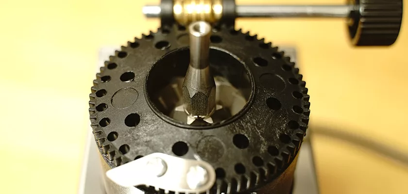 Close-up of a mechanical gear-cutting tool next to the DISCONTINUED Lelit PL043MMI Fred Espresso Coffee Grinder, showing a black circular gear and metal parts on a yellow surface. © 2025 1st-line.com