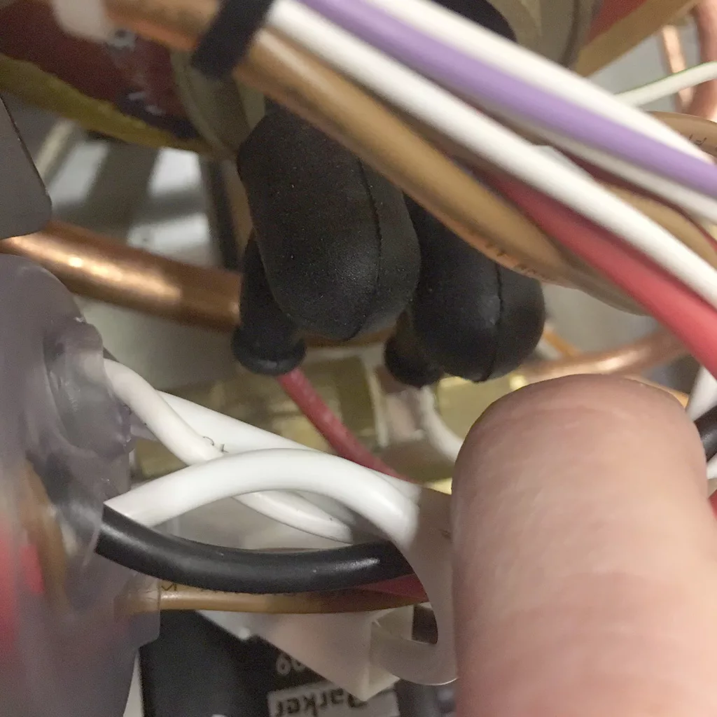 Close-up of a person’s finger touching a bundle of electrical wires—white, red, purple, and black—in a VBM Super HX espresso machine (tank only), with copper pipes and black insulated components in the background. © 2025 1st-line.com