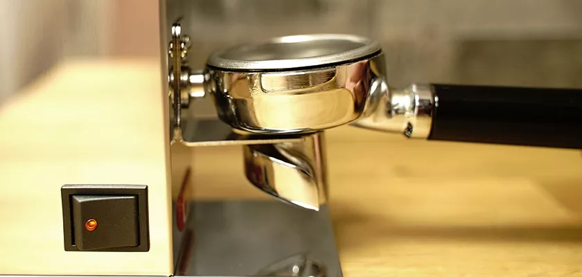 Close-up of a portafilter on an espresso machine with a black handle and orange power switch on a wooden countertop, next to the DISCONTINUED Lelit PL043MMI Fred Espresso Coffee Grinder for freshly ground coffee. © 2025 1st-line.com