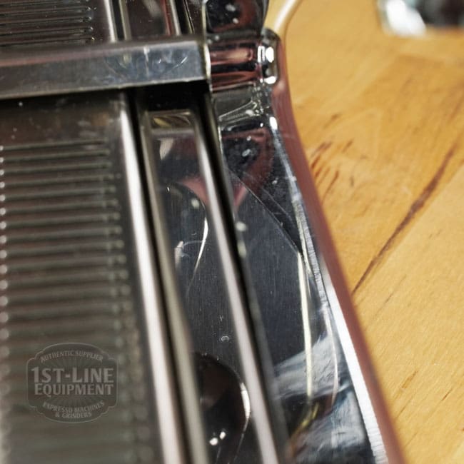 Close-up of a shiny ridged chrome part with reflections, likely from the Like NEW- La Nuova Era Cuadra Espresso Machine - V3 Chrome Color (C105), on a wooden surface. The faint 1st-Line Equipment logo is visible. © 2025 1st-line.com