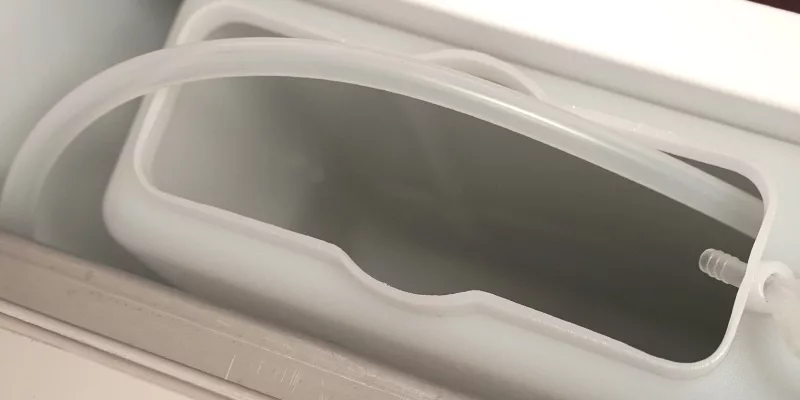 Close-up of a white plastic container with an open top, featuring a curved, transparent tube attached to a spout—likely part of the Bezzera Crema DE HX Espresso Machine water reservoir. © 2025 1st-line.com