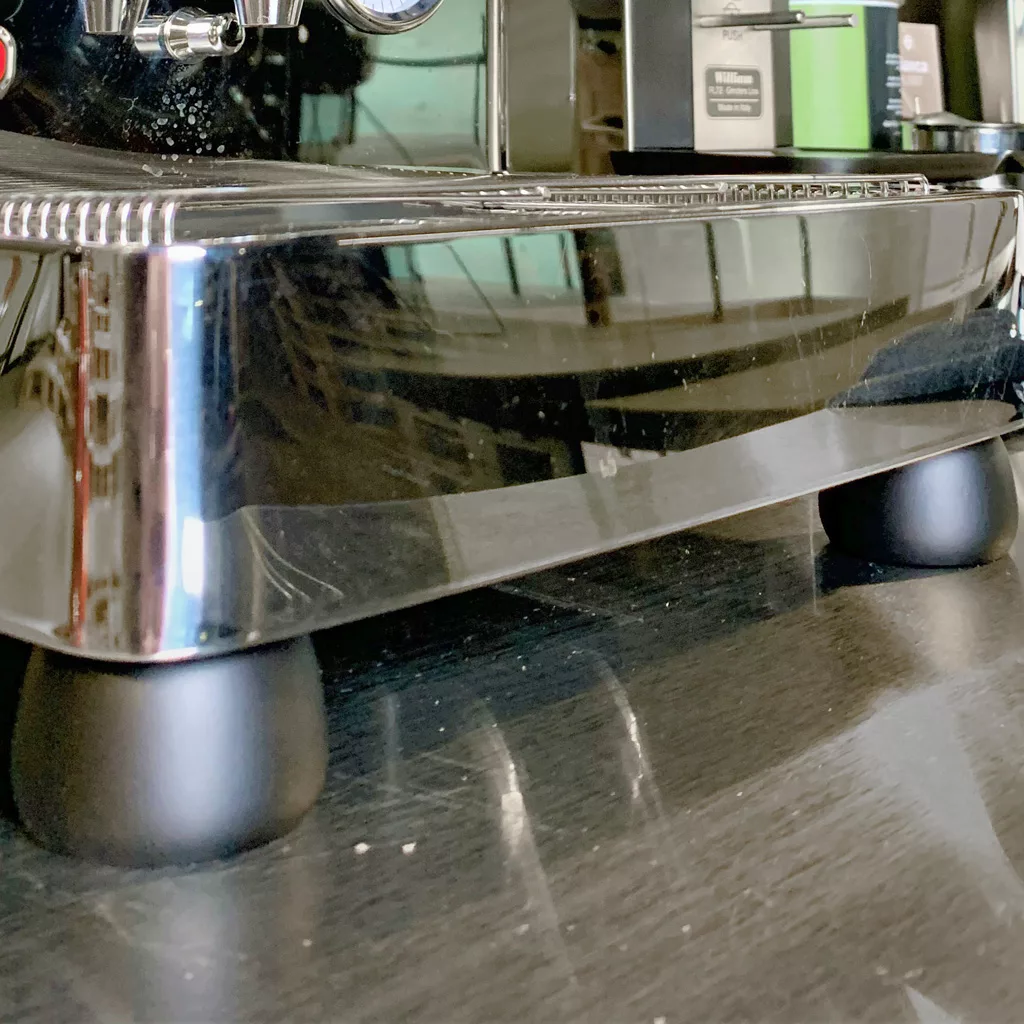 Close-up of the bottom and legs of a shiny stainless steel Lelit espresso machine on a dark countertop, with part of a coffee pod box and the Lelit PLA2201 Black Wood Kit visible in the background. © 2025 1st-line.com