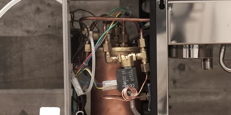 Close-up of the internal components of a Bezzera Crema DE HX Espresso Machine, showcasing HX copper pipes, metal valves, electrical wires, and connectors set against a blurred background. © 2025 1st-line.com