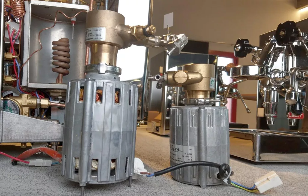 Two rotary vane pumps with silver metal housings and brass fittings, rated at 120 watts and 150 watts, are placed on a carpeted floor. Coffee machine components and equipment are visible in the background. © 2025 1st-line.com