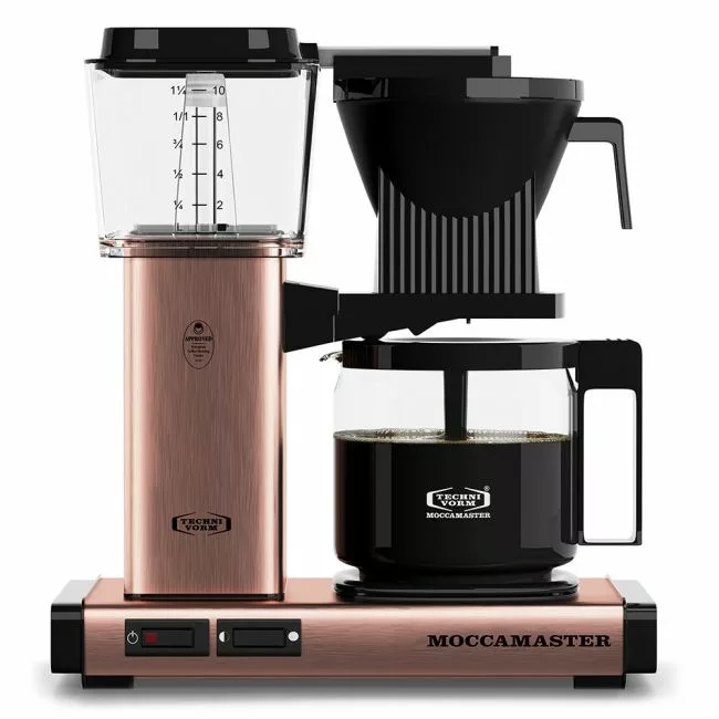 A Technivorm Moccamaster coffee brewer with a copper finish, featuring a KBG glass carafe, black handle, water reservoir with measurement markings, and control switches on the front base. © 2025 1st-line.com
