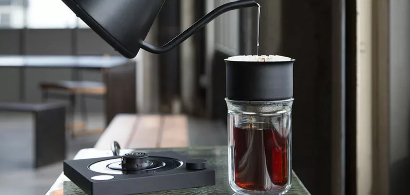 A DISCONTINUED Fellow Stagg EKG Electric Kettle - 0.9L, 120V in black pours hot water over coffee grounds in a dripper on a glass jar, with its electric base nearby in a modern, sunlit room. © 2025 1st-line.com