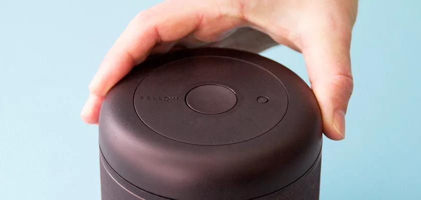 A hand presses down on the lid of a DISCONTINUED - Fellow Atmos Vacuum Canister 1.2 L - Stainless Steel, set against a light blue background. © 2025 1st-line.com