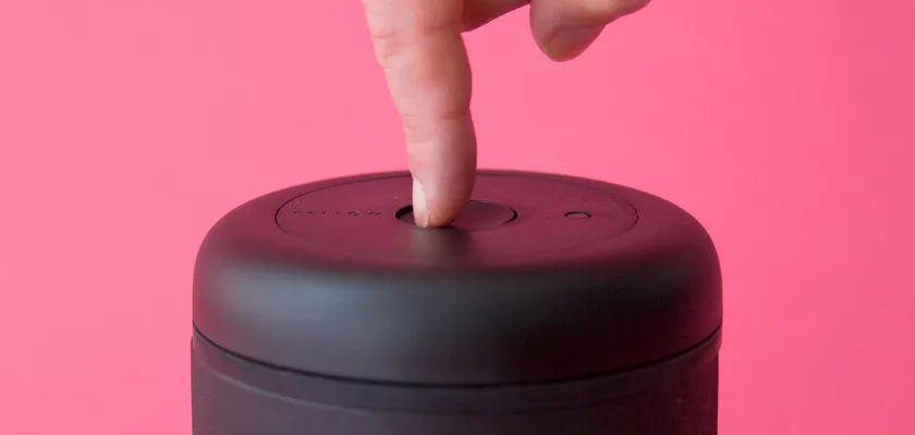 A hand presses the power button on a DISCONTINUED - Fellow Atmos Vacuum Canister 1.2 L - Stainless Steel, set against a pink background. © 2025 1st-line.com