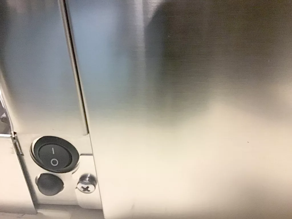 A close-up of a metallic surface featuring a round black power switch set to off, a small round button, and a screw—possibly part of an appliance like the Lelit Mara X or its water tank connection, ideal for repair scenarios. © 2025 1st-line.com