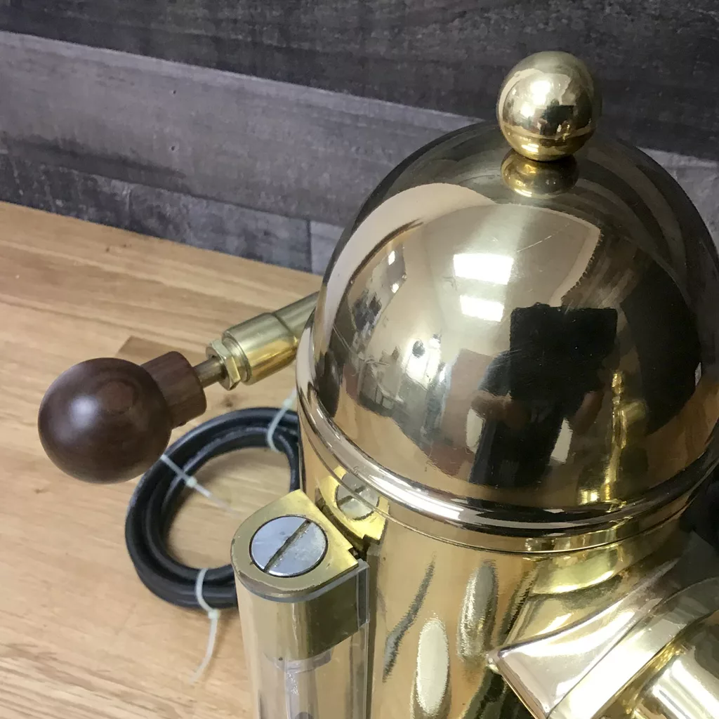 Close-up of a SOLD IN STORE PICKUP ONLY USED La Pavoni 16-Cup Professional Brass Lever Espresso Machine, featuring a dome top, wooden handle, and black cord, set on a wooden surface with a gray wood wall backdrop. © 2025 1st-line.com