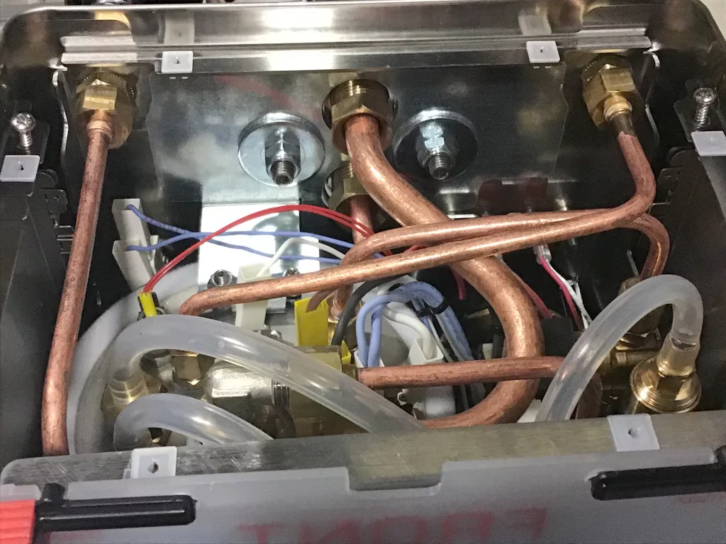 Close-up of the inside of a Lelit Mara X machine showing copper pipes, white and blue wires, and clear plastic tubing connected to various metal fittings, highlighting the water tank connection for repair or maintenance. © 2025 1st-line.com