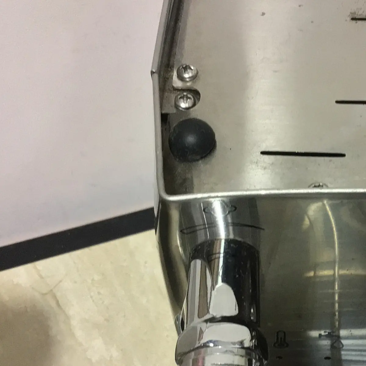 Close-up of the corner of a used SOLD SIDEWALK SALE - Lelit Mara X PL62X HX Espresso Machine (E213), featuring screws, a black rubber foot, and part of a shiny metal handle against a light floor and wall background.