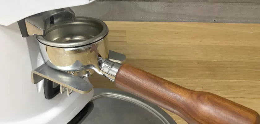 A close-up of a portafilter with a wooden handle attached to a white espresso machine, set on a wooden countertop beside a DISCONTINUED Fiorenzato F4 Nano V2 Espresso Coffee Grinder. © 2025 1st-line.com