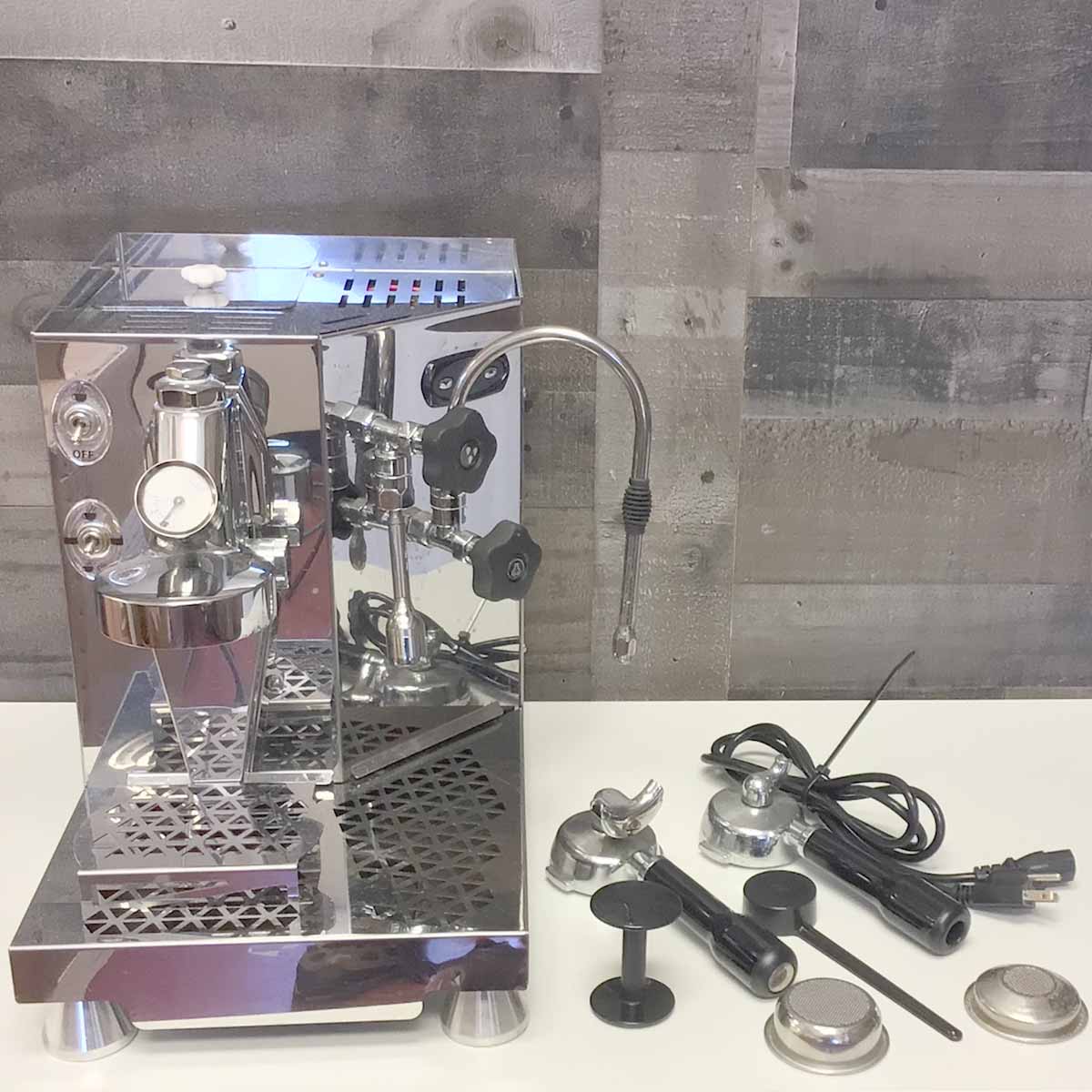 A returned & refurbished ACS Minima Dual Boiler Espresso Machine from our Sidewalk Sale sits on a counter with a portafilter, tamper, baskets, and power cord, set against a gray wood-paneled wall background. © 2025 1st-line.com