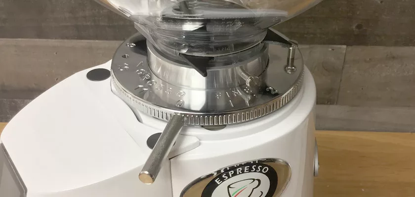 Close-up of a DISCONTINUED Fiorenzato F4 Nano V2 Espresso Coffee Grinder featuring a clear hopper, silver adjustment dial, and lever with the Espresso label on the front, shown on a wooden surface with a wooden wall background. © 2025 1st-line.com