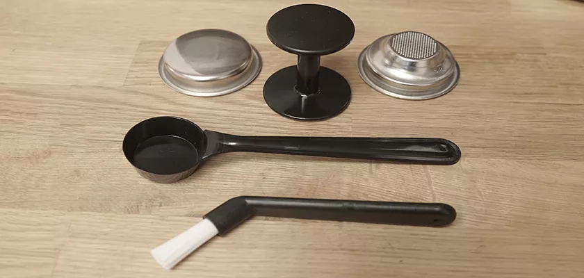 A portafilter basket, coffee tamper, black measuring scoop, and white-bristle cleaning brush on wood—essential accessories for your Bezzera New Hobby Espresso Machine or any espresso setup. © 2025 1st-line.com