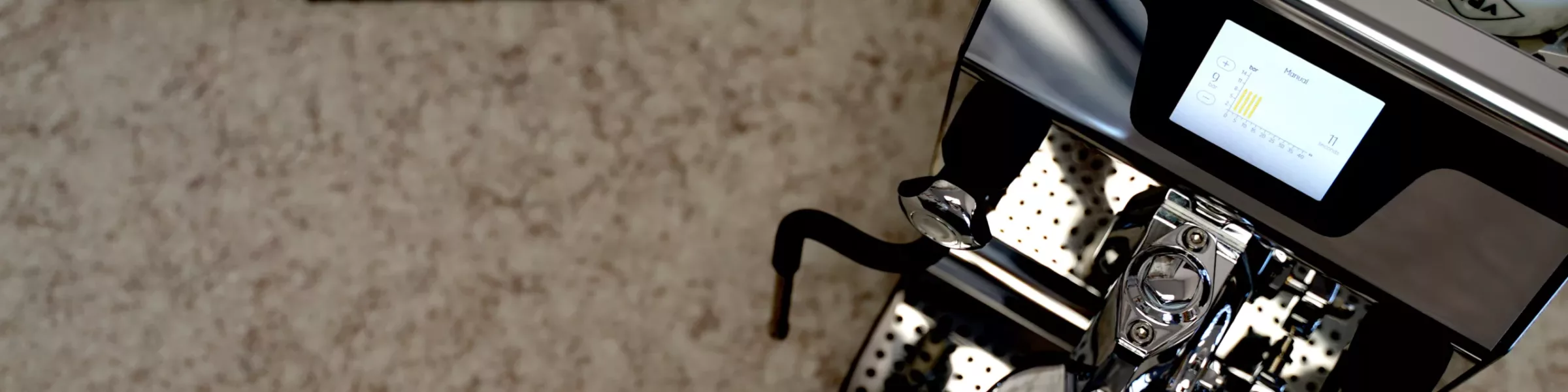 Close-up of a modern dual boiler espresso machine with a digital display screen and metal drip tray, set against a speckled countertop background. Only part of the machine is visible in the frame. © 2025 1st-line.com