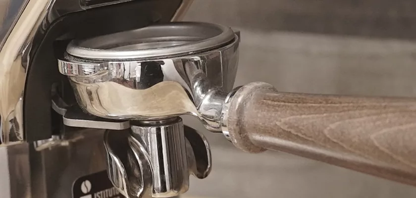 A portafilter with a wooden handle is attached to an espresso machine, ready for brewing, next to a DISCONTINUED Eureka Olympus 75E High Speed Espresso Coffee Grinder with titanium burrs for precise grounds. © 2025 1st-line.com