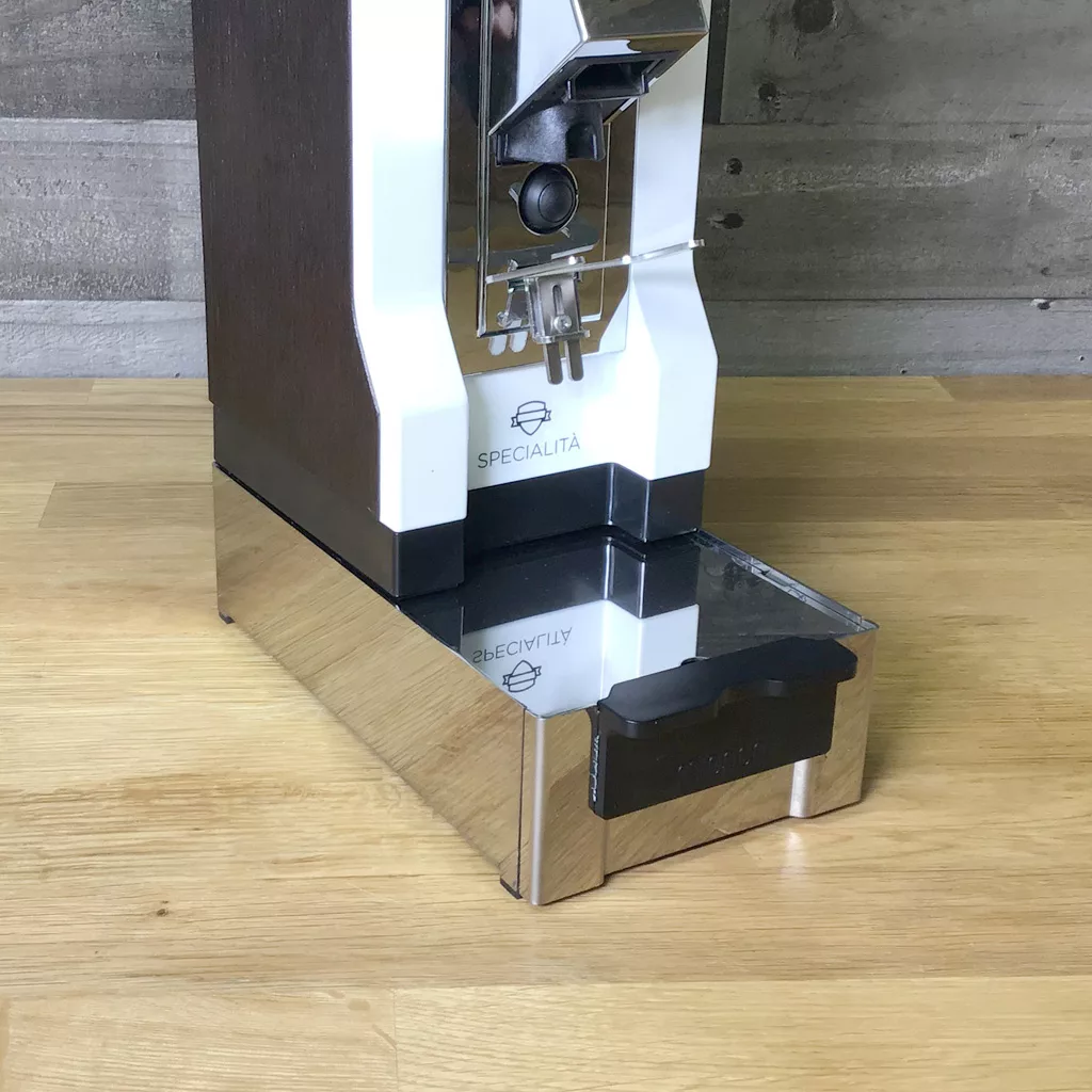 A close-up of a Eureka 2311.0119A0R01-901 Mignon Base/Knockbox placed on a wooden surface, with a concrete wall in the background. © 2025 1st-line.com