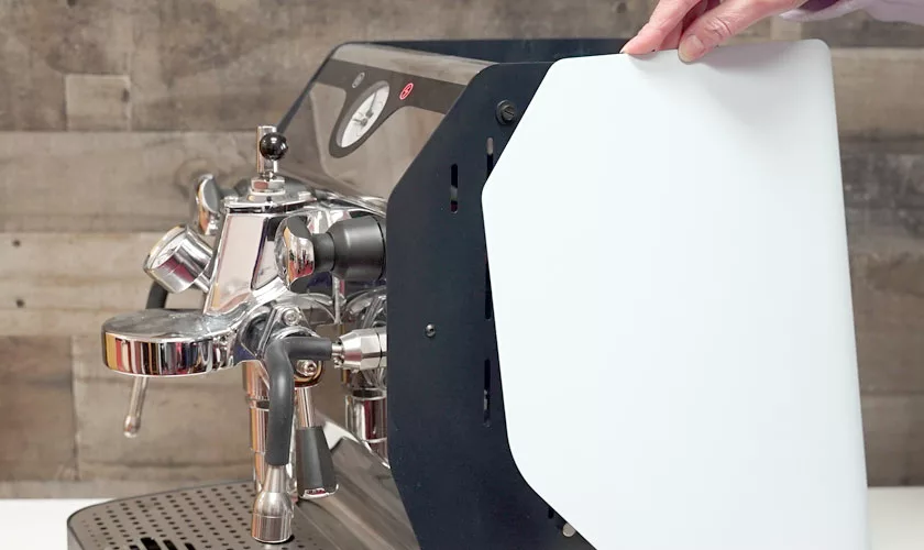 A hand holds a white side panel of the VBM Domobar Super Analogic HX Espresso Machine with Flow Control, featuring multiple levers and handles, set on a counter with a wooden wall in the background. © 2025 1st-line.com