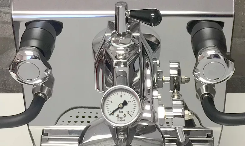 Close-up of the VBM Junior Digital HX Espresso Machine w/ Flow Control, showcasing a central pressure gauge marked with bar Coffee Sensor and two symmetrical portafilter holders that reflect on the metallic surface. © 2025 1st-line.com