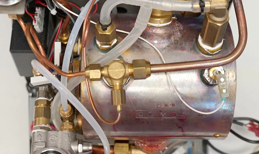Close-up view of the internal components of a VBM Domobar Super Analogic HX Espresso Machine w/ Flow Control, featuring a metal boiler with brass fittings, copper pipes, flow control parts, white plastic tubes, and electrical wiring. © 2025 1st-line.com