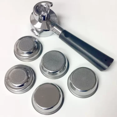 A Lelit PLA572V 57 mm Coffee Slide Spout VIP Handle Portafilter with a black handle sits on a white surface alongside five metal filter baskets, including both fine mesh and solid bottom types. © 2025 1st-line.com